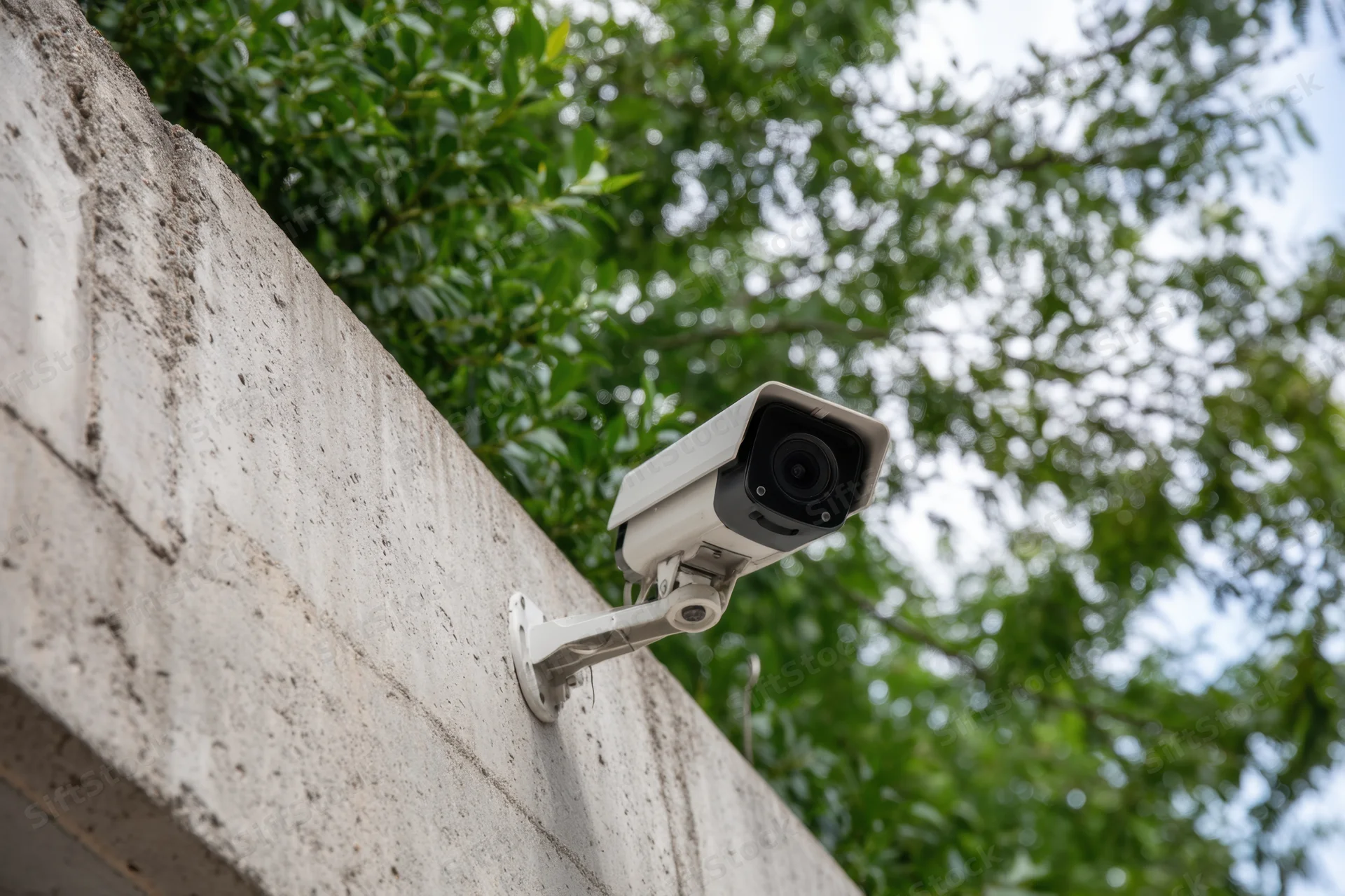 Surveillance camera mounted on textured concrete wall with green foliage in background