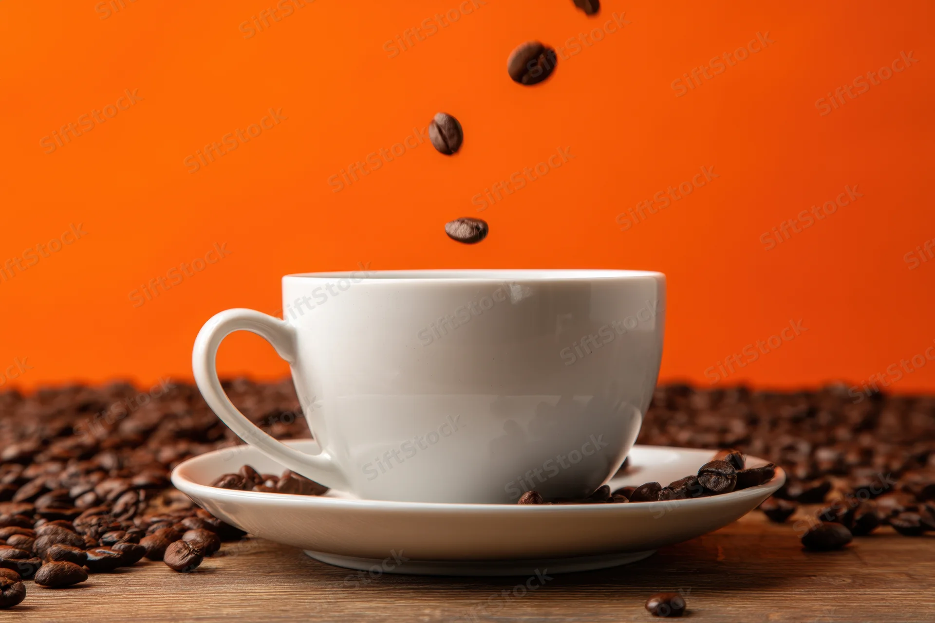 White ceramic coffee cup with saucer on wooden table surrounded by falling coffee beans against orange background