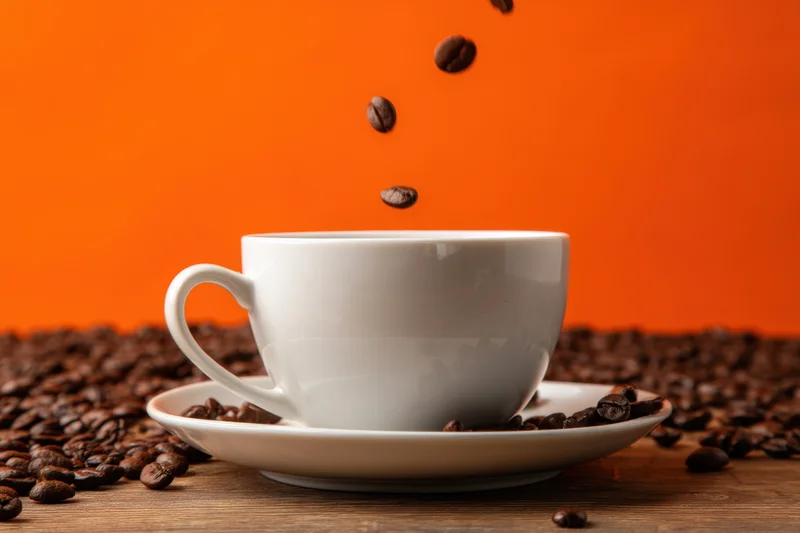 White ceramic coffee cup with saucer on wooden table surrounded by falling coffee beans against orange background