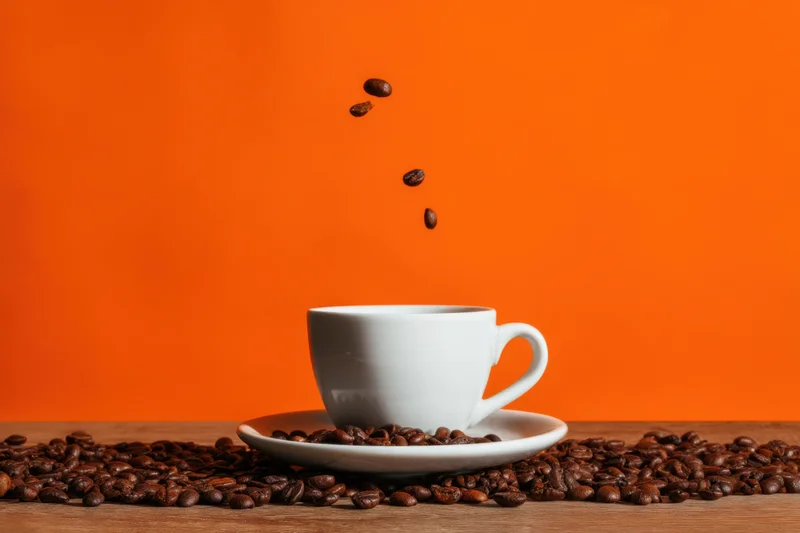 White ceramic coffee cup with saucer on wooden table surrounded by roasted coffee beans with beans falling into cup against orange background