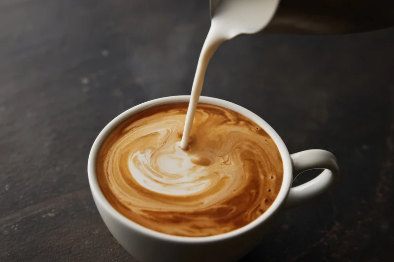 Milk being poured into a steaming latte in a white ceramic cup on a dark surface