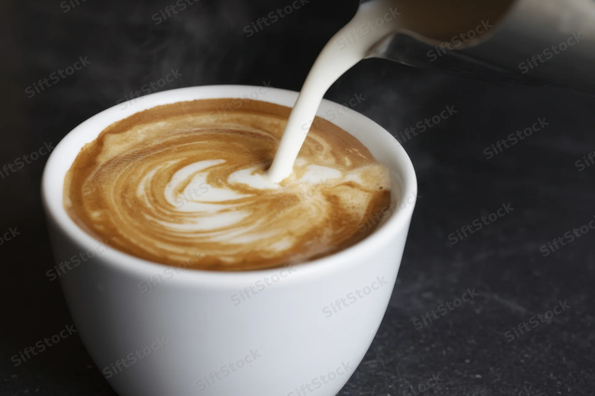 Milk being poured into a steaming latte in a white ceramic cup on a dark surface