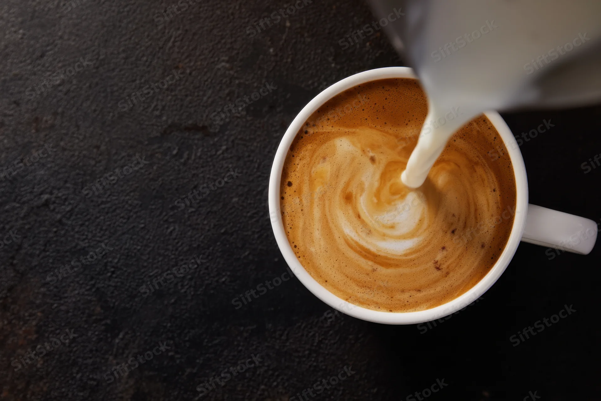 Milk being poured into a white ceramic coffee cup creating a swirling foam pattern on a dark surface