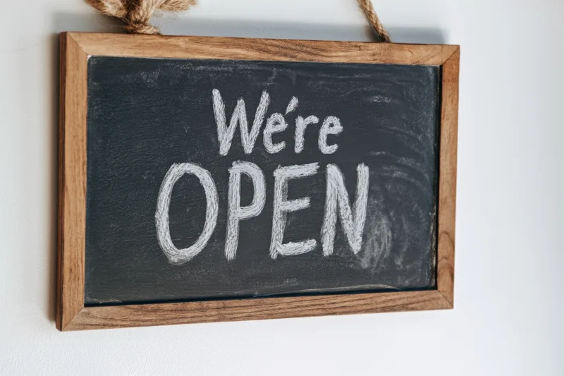 Wooden-framed chalkboard sign with white chalk text reading we're open hanging on a white wall by rope