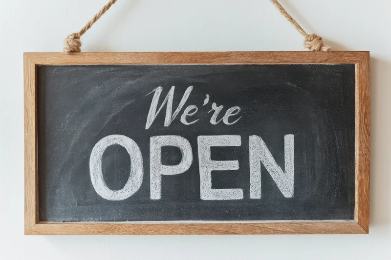 Wooden-framed chalkboard sign with white chalk text reading we're open hanging on a white wall