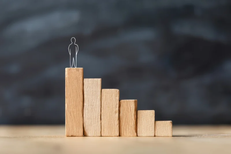 Wireframe figure standing atop wooden blocks arranged in ascending staircase on wooden surface