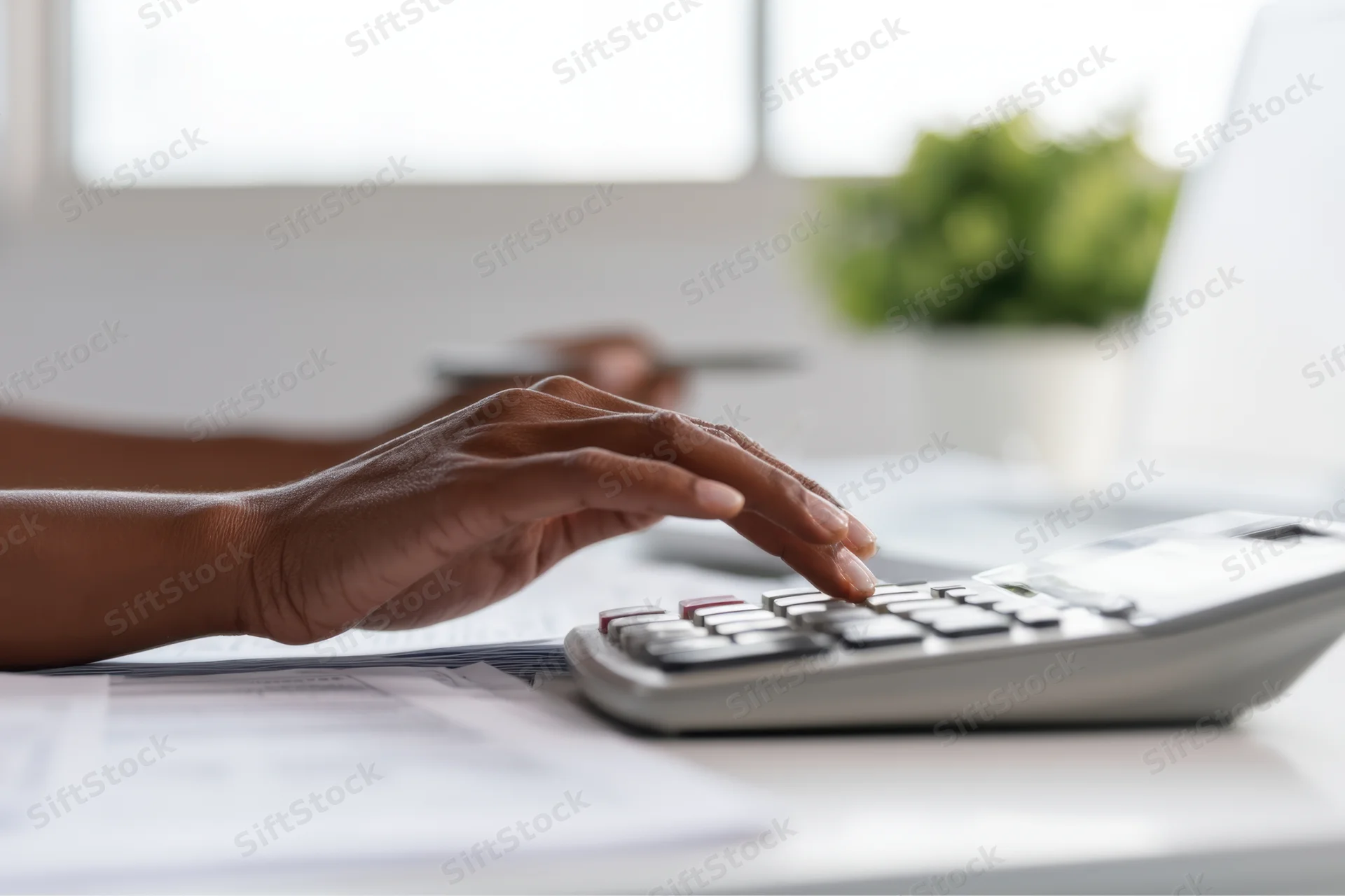 Woman’s hands using calculator on office desk with documents and laptop nearby