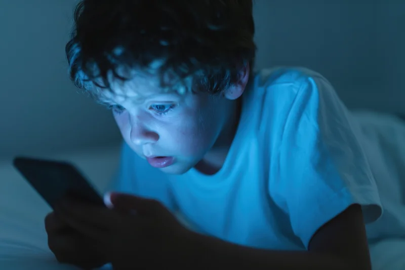 Young boy lying on bed focused on smartphone screen in dark room