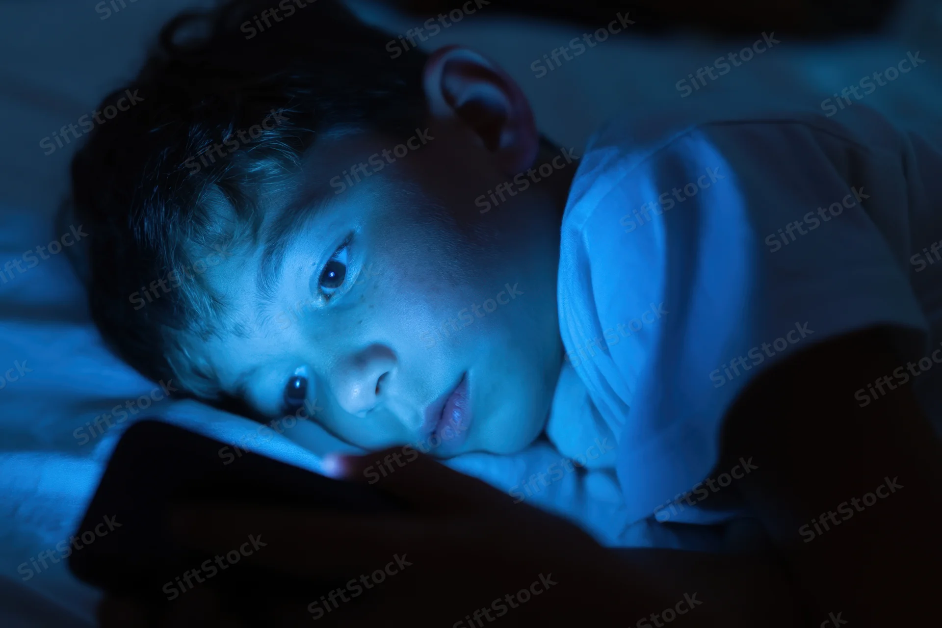 Young boy lying on bed focused on smartphone screen at night