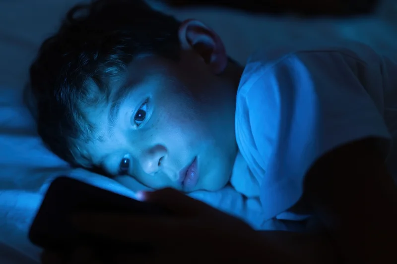 Young boy lying on bed focused on smartphone screen at night