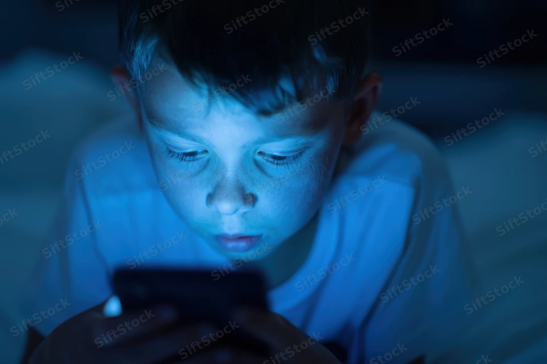 Young boy lying on bed focused on smartphone screen in dark room