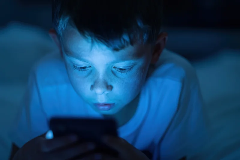 Young boy lying on bed focused on smartphone screen in dark room