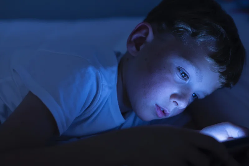 Young boy lying on bed focused on smartphone screen in dark bedroom
