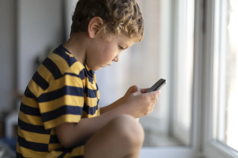 Young boy sitting on windowsill using smartphone indoors
