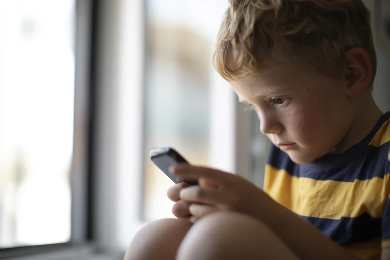 Young boy sitting on windowsill holding smartphone and looking at screen in focus
