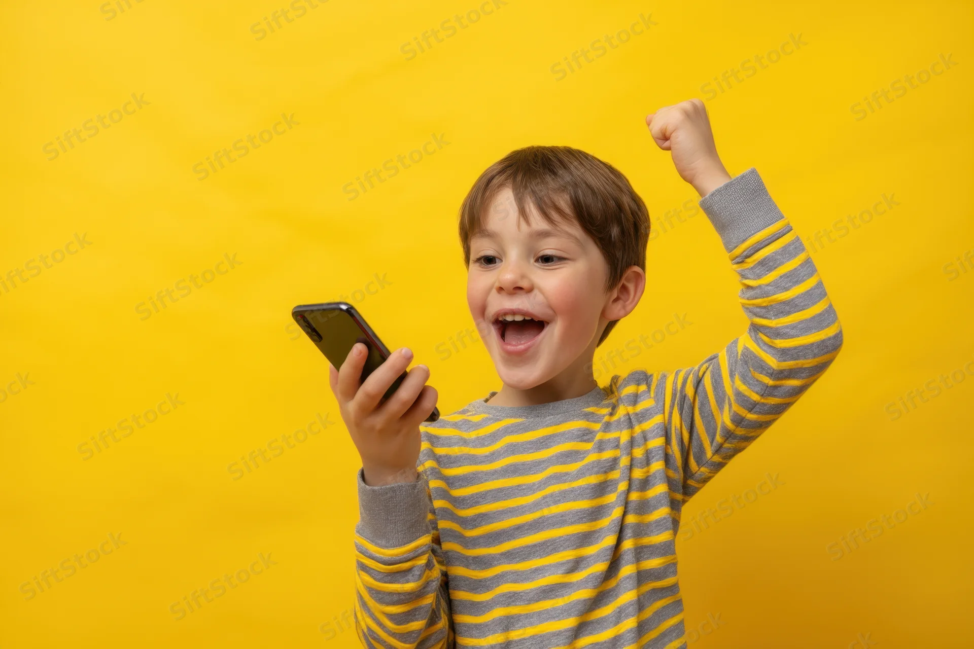 Young boy holding smartphone and raising fist in celebration against yellow background