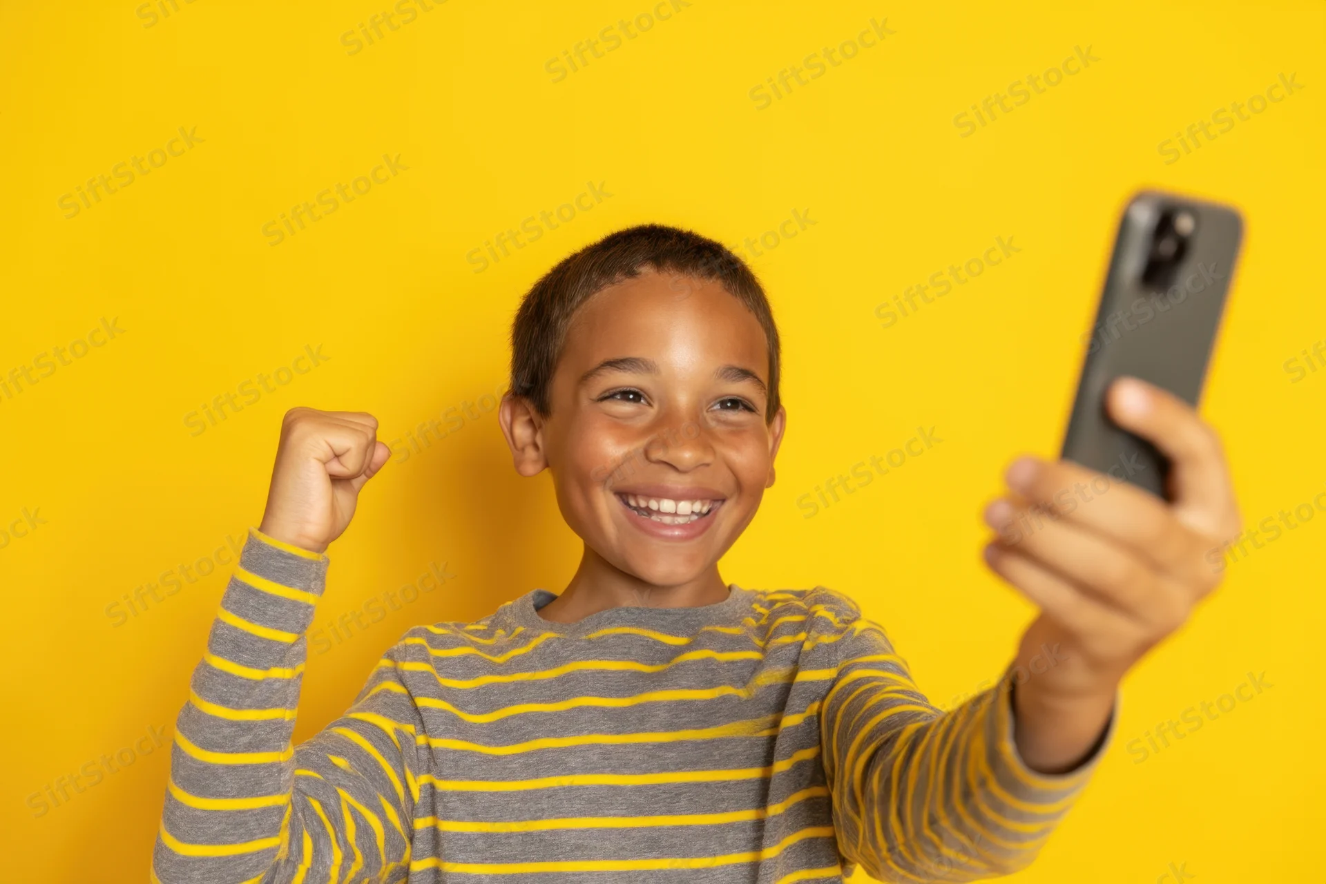 Young boy smiling and taking a selfie with smartphone against yellow background, raising fist in excitement