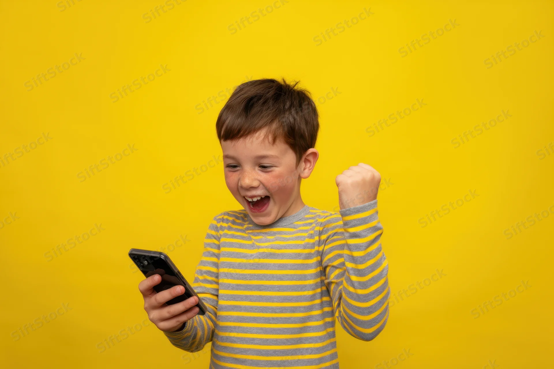 Young boy laughing and raising fist while holding smartphone against yellow background