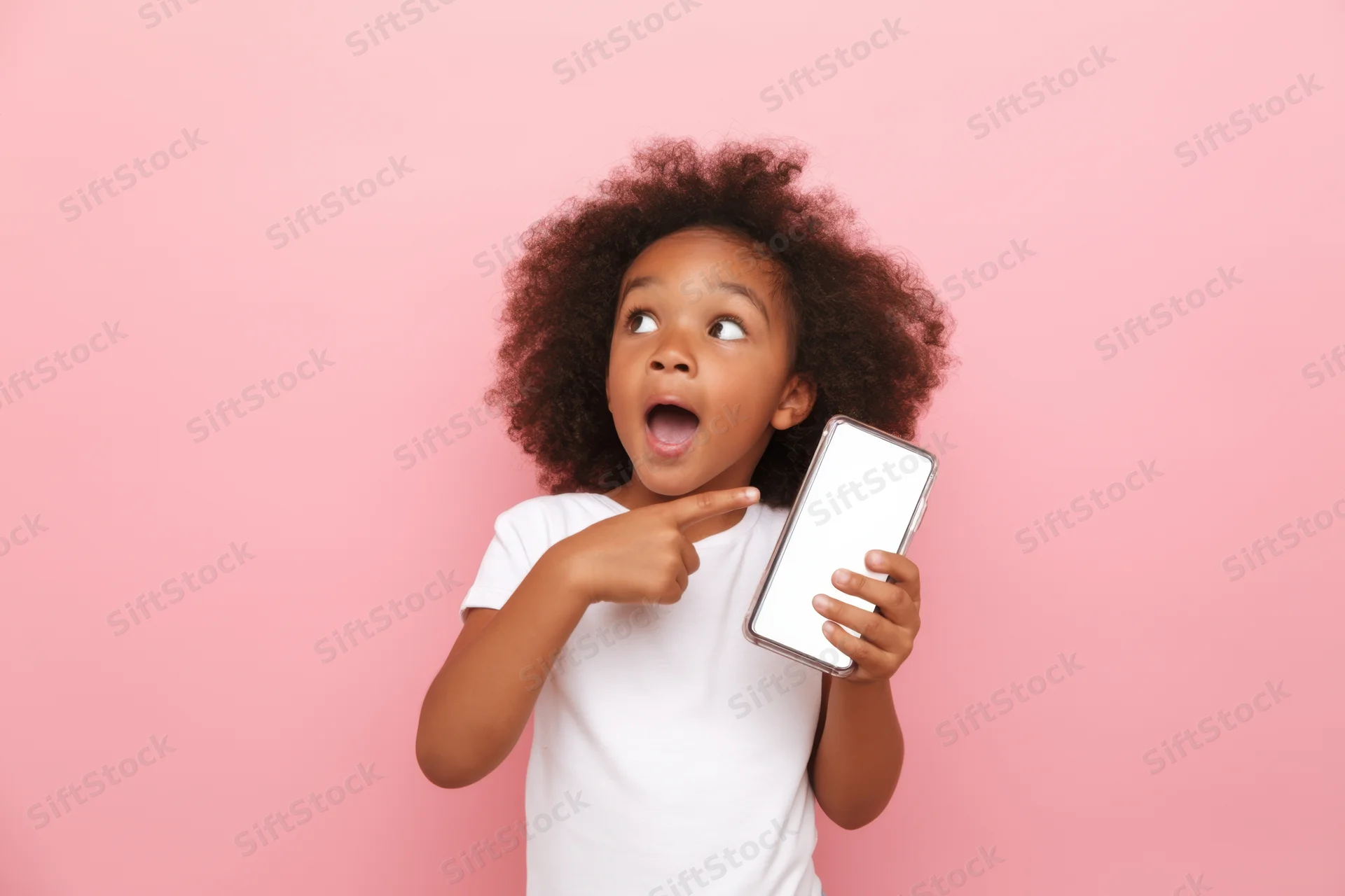 Young black girl expressing surprise while pointing at smartphone screen against pink background