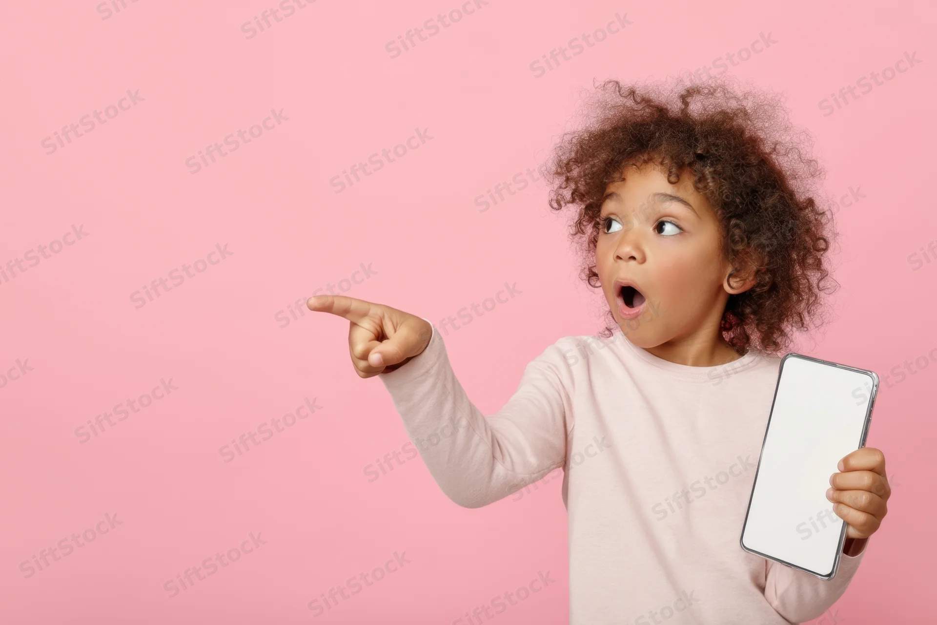 Young girl with curly hair pointing excitedly while holding a blank smartphone screen against a pink background