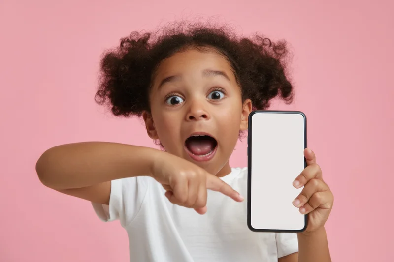Young black girl expressing excitement while pointing at a blank smartphone screen held in her hand against a pink background