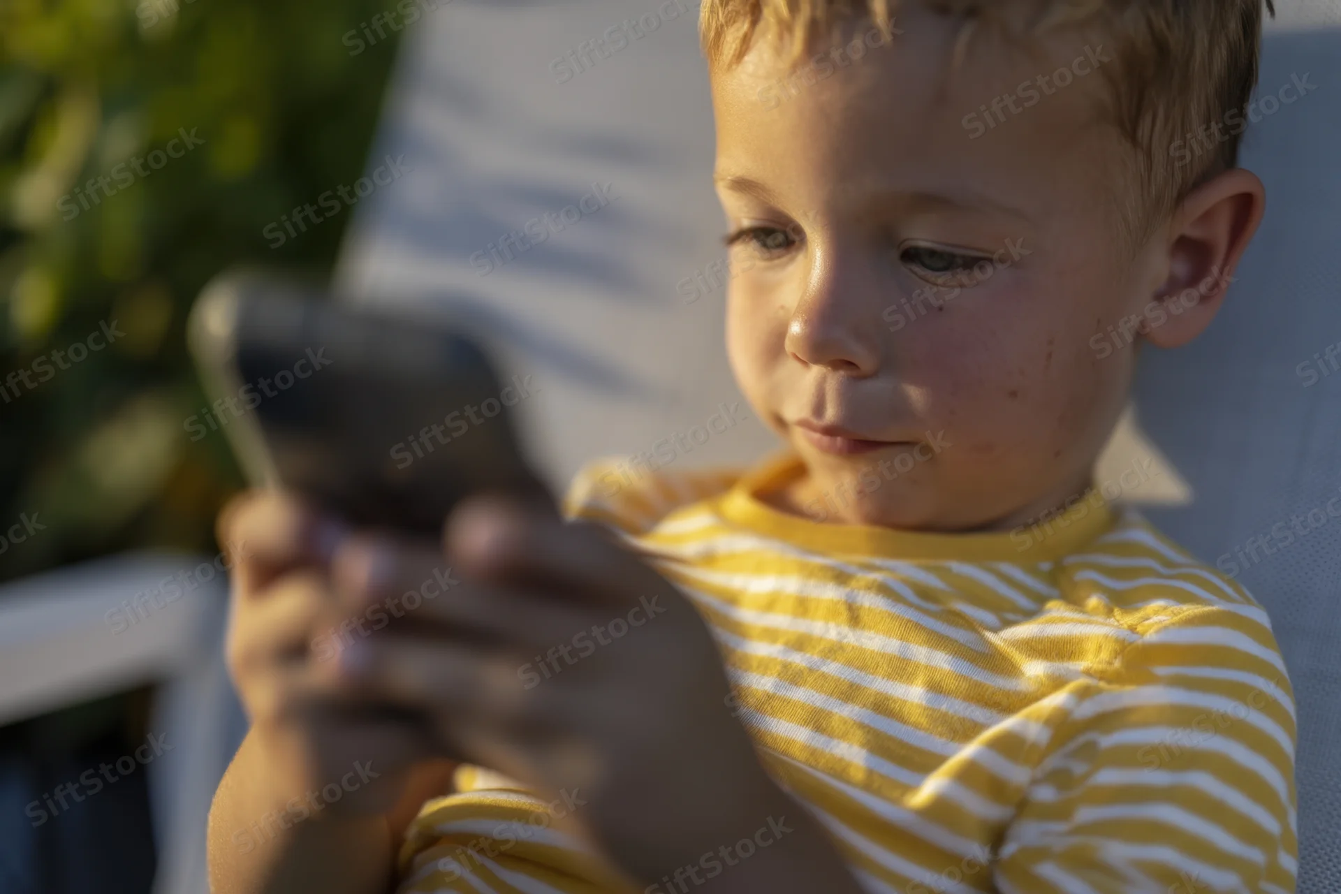 Young boy focused on smartphone outdoors in garden setting wearing yellow striped shirt