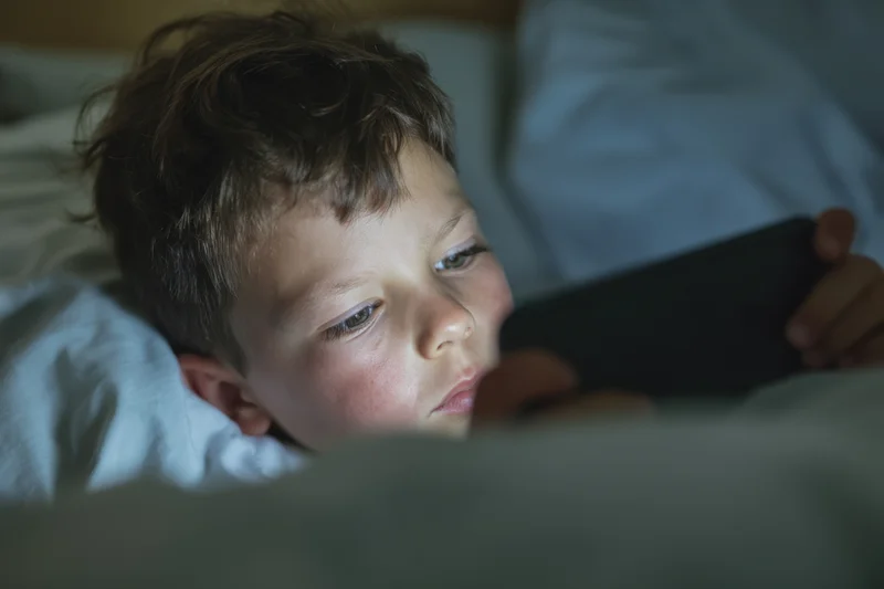 Young boy lying in bed focused on smartphone screen at bedtime