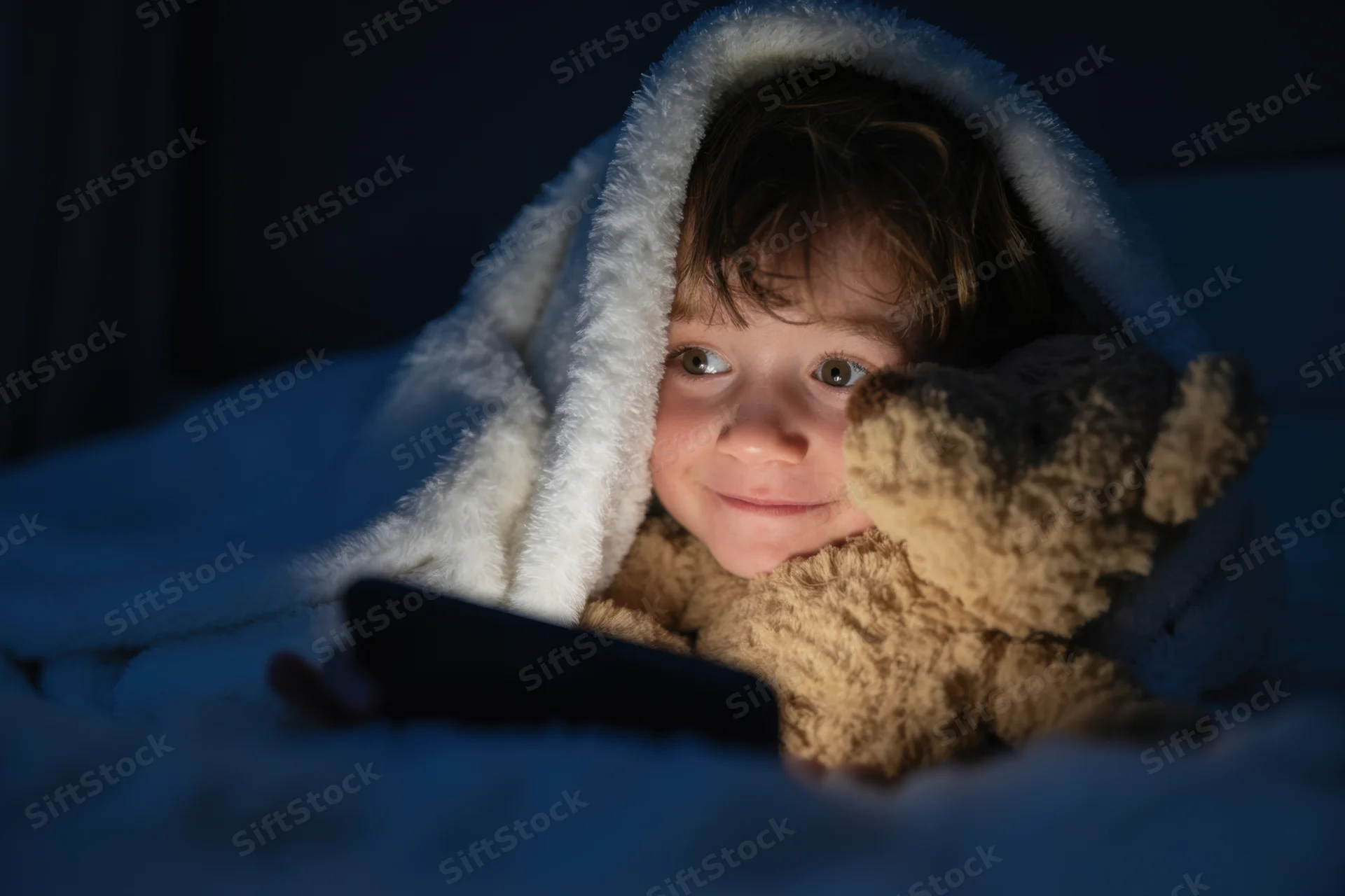 Young boy under blanket with teddy bear and smartphone in bed at night