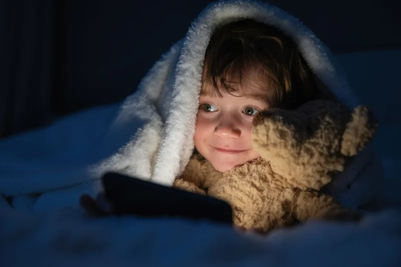 Young boy under blanket with teddy bear and smartphone in bed at night