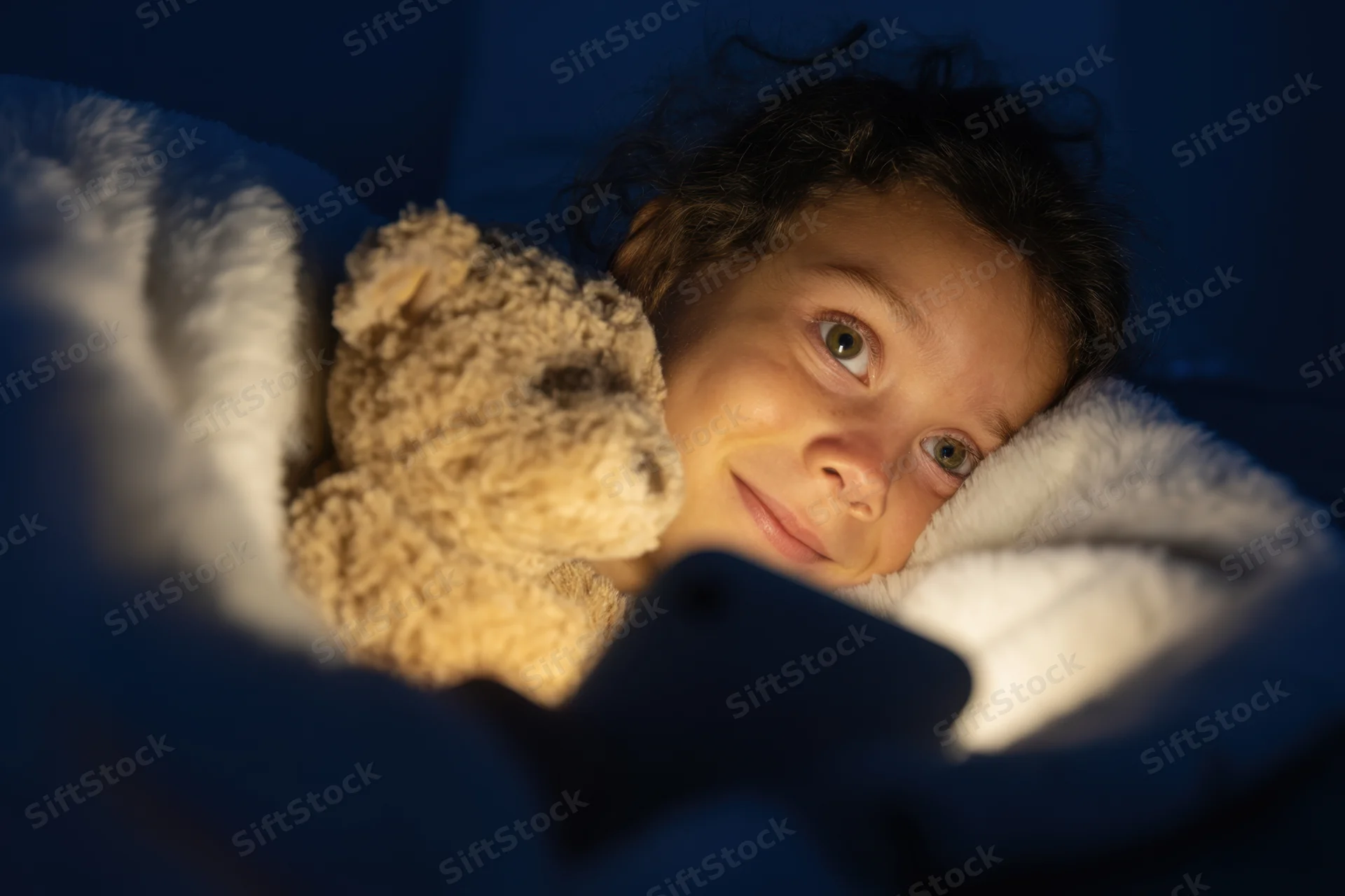 Young girl lying under a blanket with a teddy bear, illuminated by phone screen light at night