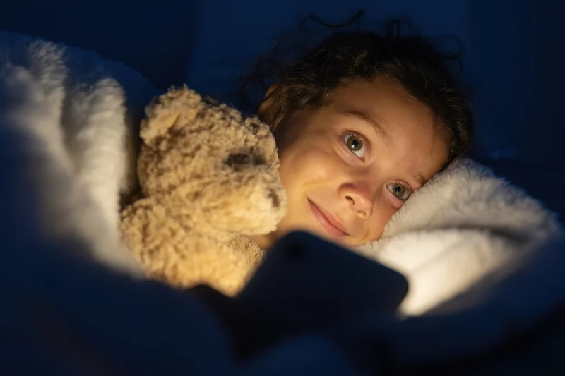 Young girl lying under a blanket with a teddy bear, illuminated by phone screen light at night