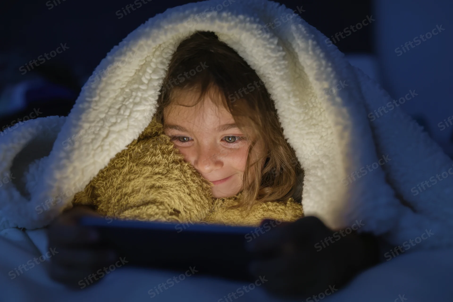 Young girl under blanket holding tablet and smiling in bed at night
