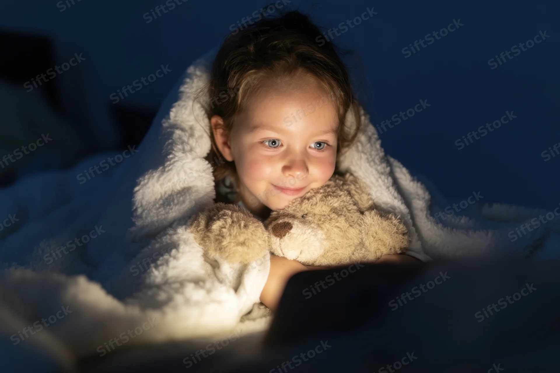 Young girl smiling under blanket with teddy bear in dimly lit bedroom at night