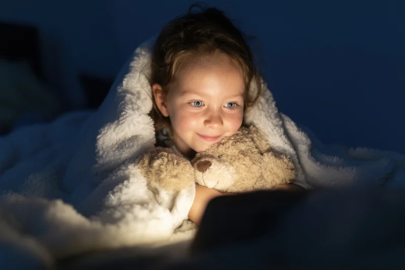 Young girl smiling under blanket with teddy bear in dimly lit bedroom at night