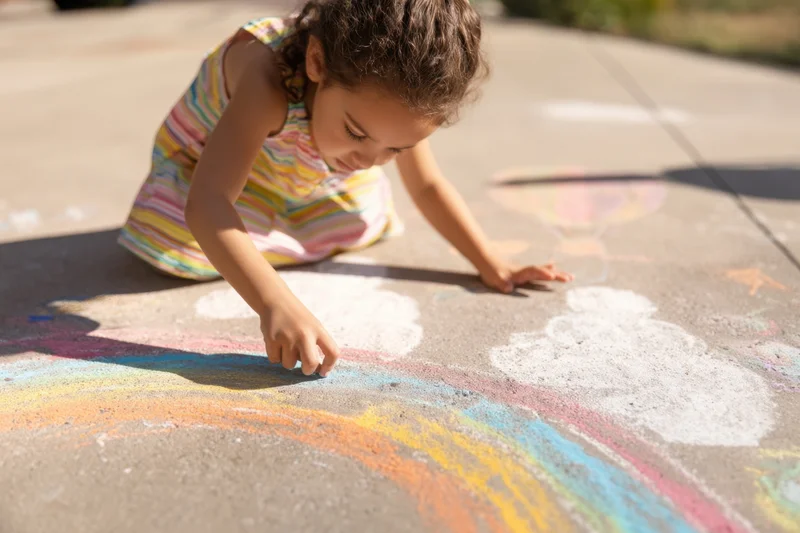 Young girl drawing a rainbow with chalk on a concrete sidewalk outdoors
