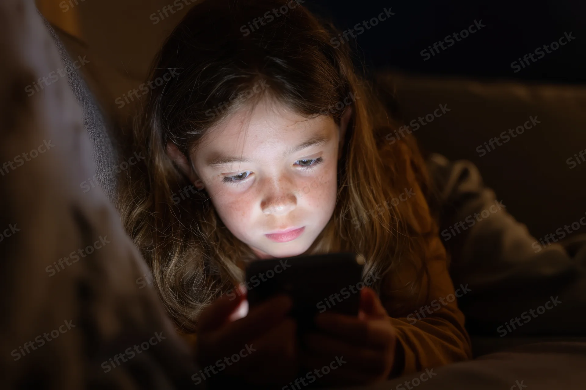 Young girl lying on couch focused on smartphone screen in dim indoor setting