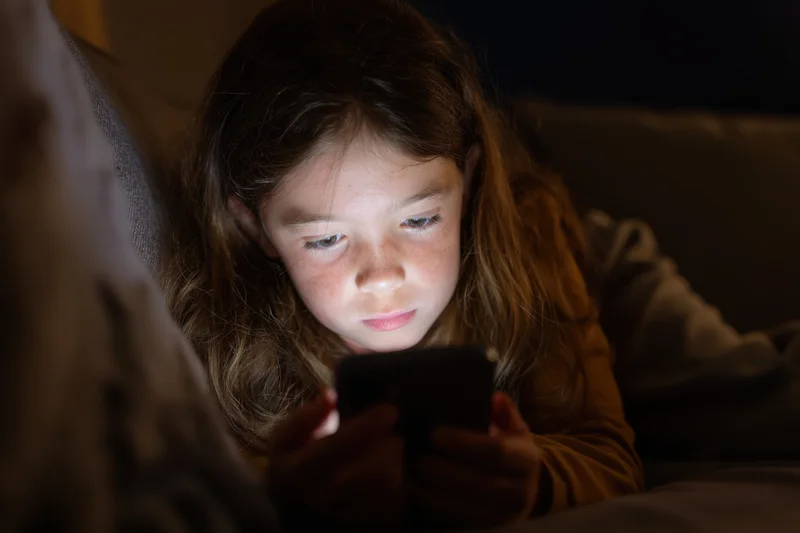 Young girl lying on couch focused on smartphone screen in dim indoor setting