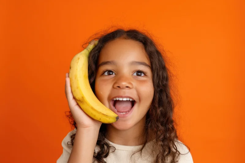 Young girl smiling widely while holding a banana up to her face against an orange background