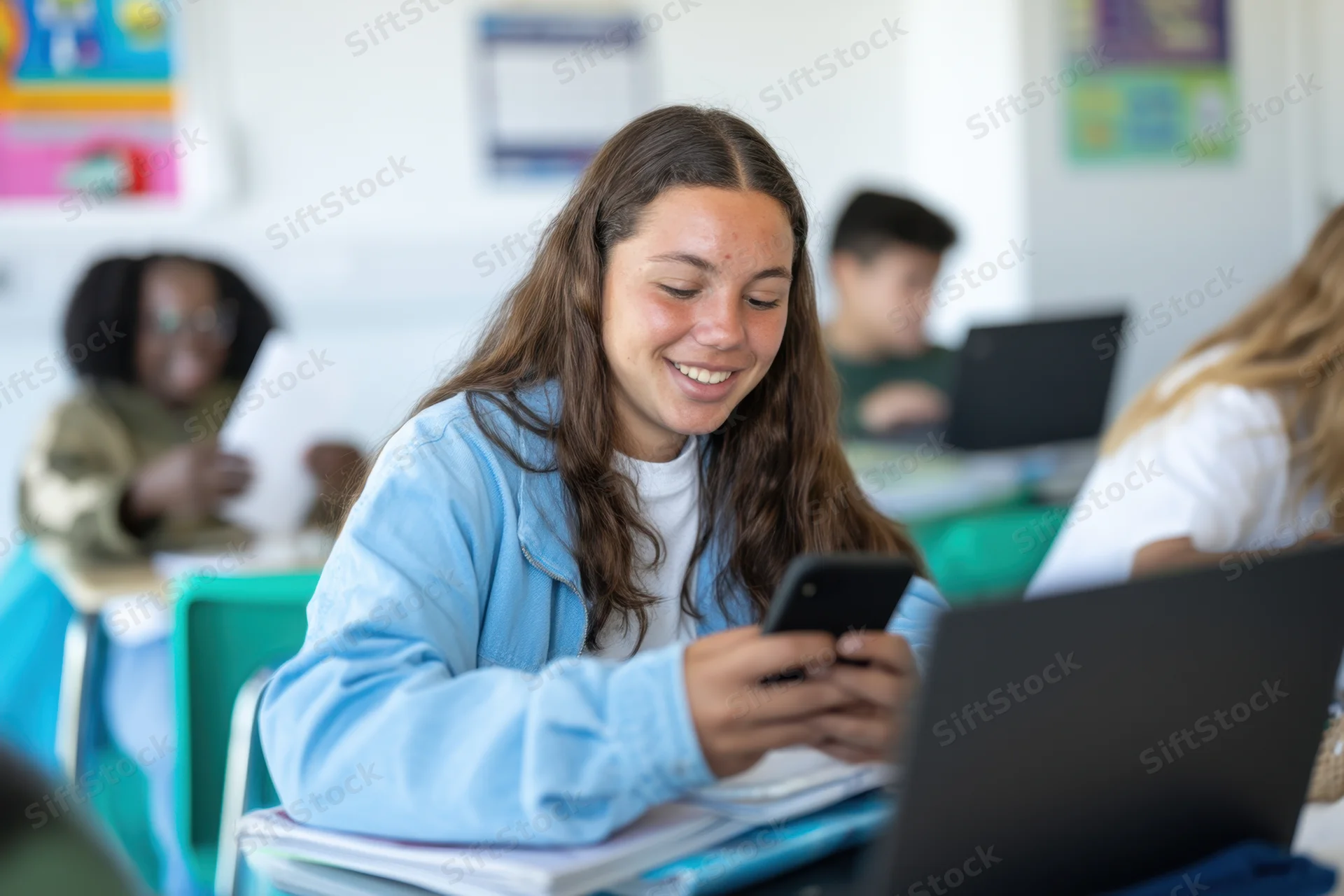 Young girl smiling while using smartphone at desk in classroom with classmates studying around her