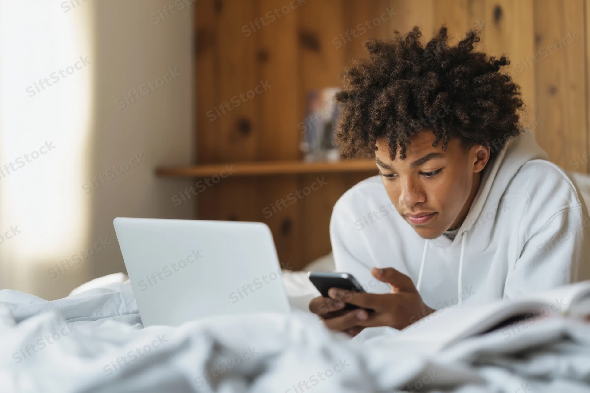 Young black male adolescent lying on bed focused on smartphone with laptop nearby