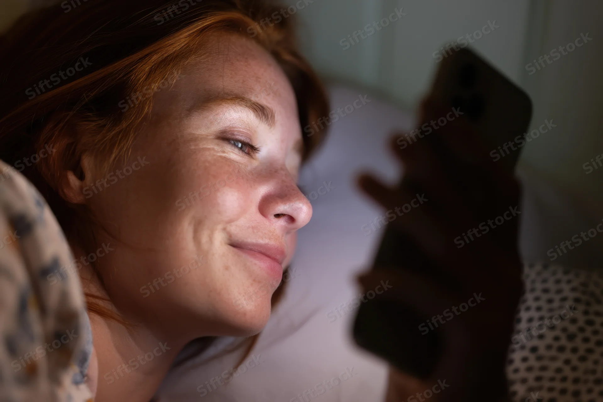 Young woman lying in bed smiling while holding smartphone close to face at night