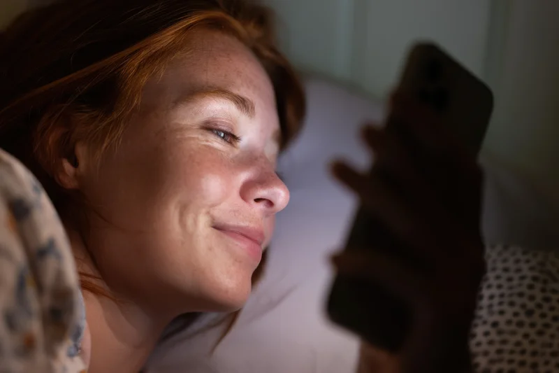 Young woman lying in bed smiling while holding smartphone close to face at night