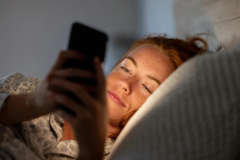 Young woman lying in bed holding smartphone close to her face at night