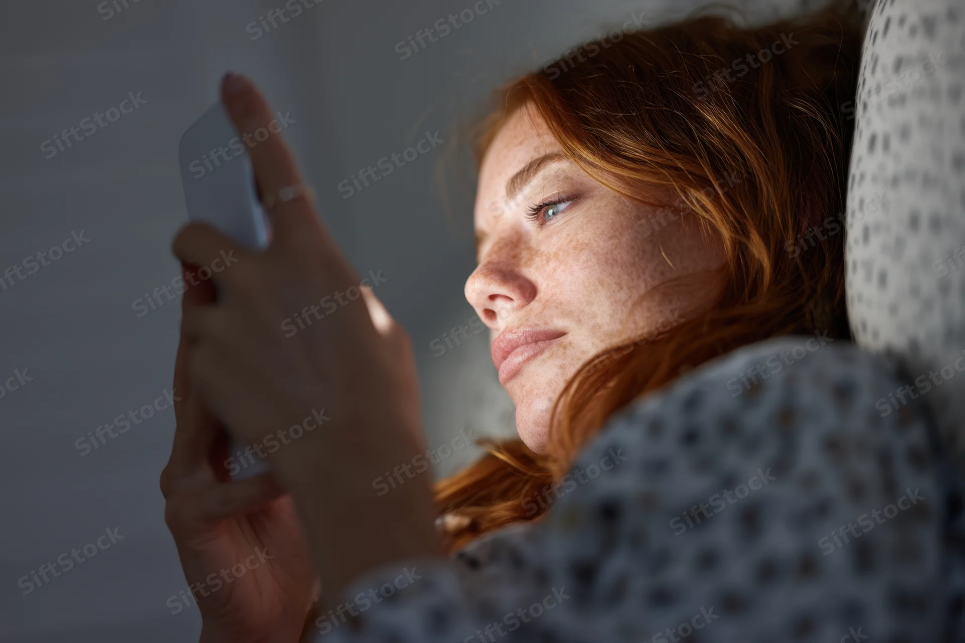 Woman lying in bed holding smartphone close to her face in dim light