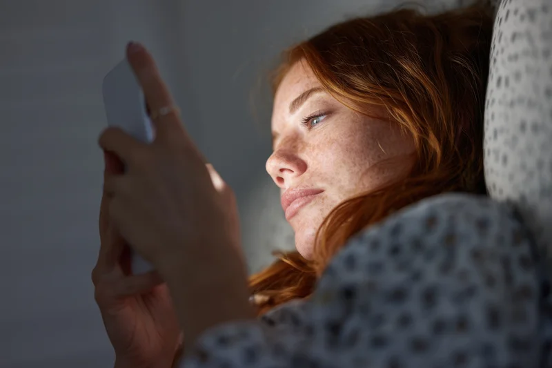 Woman lying in bed holding smartphone close to her face in dim light