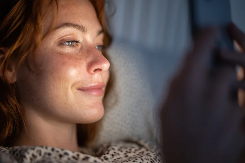 Woman lying in bed holding smartphone close to her face, smiling in dim light