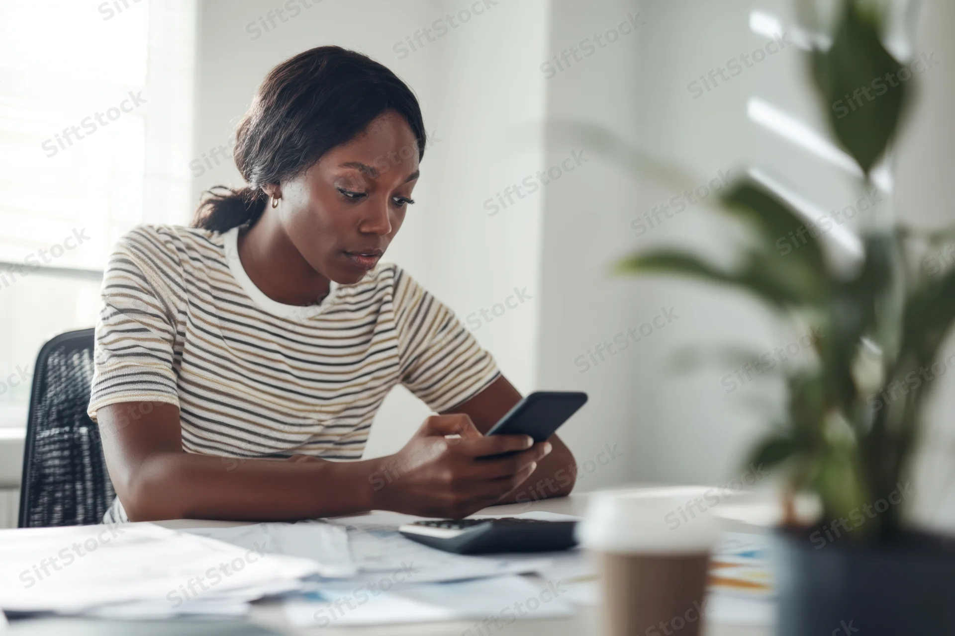 Young black woman focused on smartphone at modern desk with documents and coffee