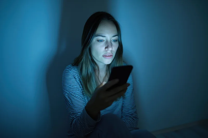 Young woman sitting on floor in dim room holding smartphone, illuminated by screen light