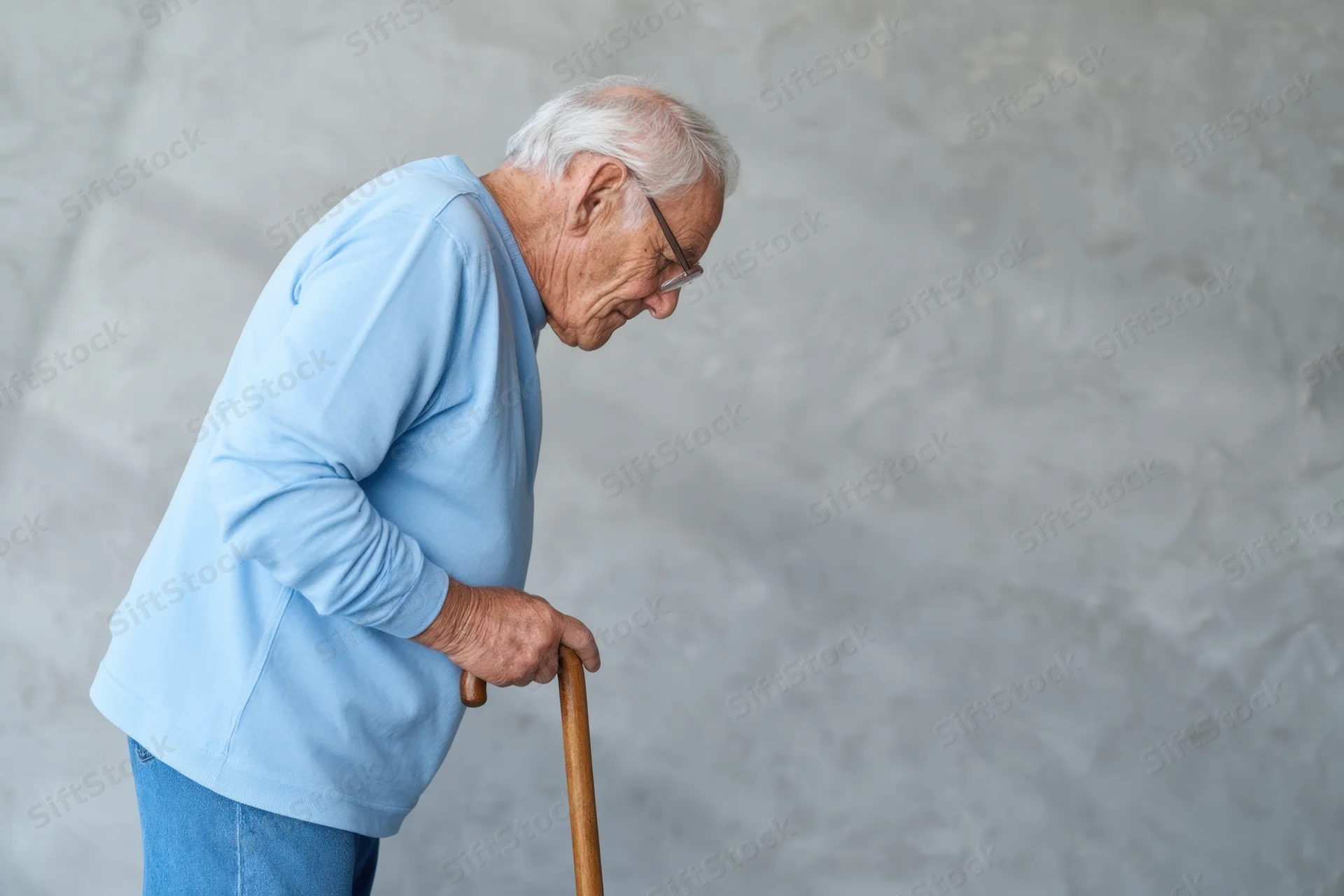 Elderly man using wooden cane while walking with bent posture against plain wall