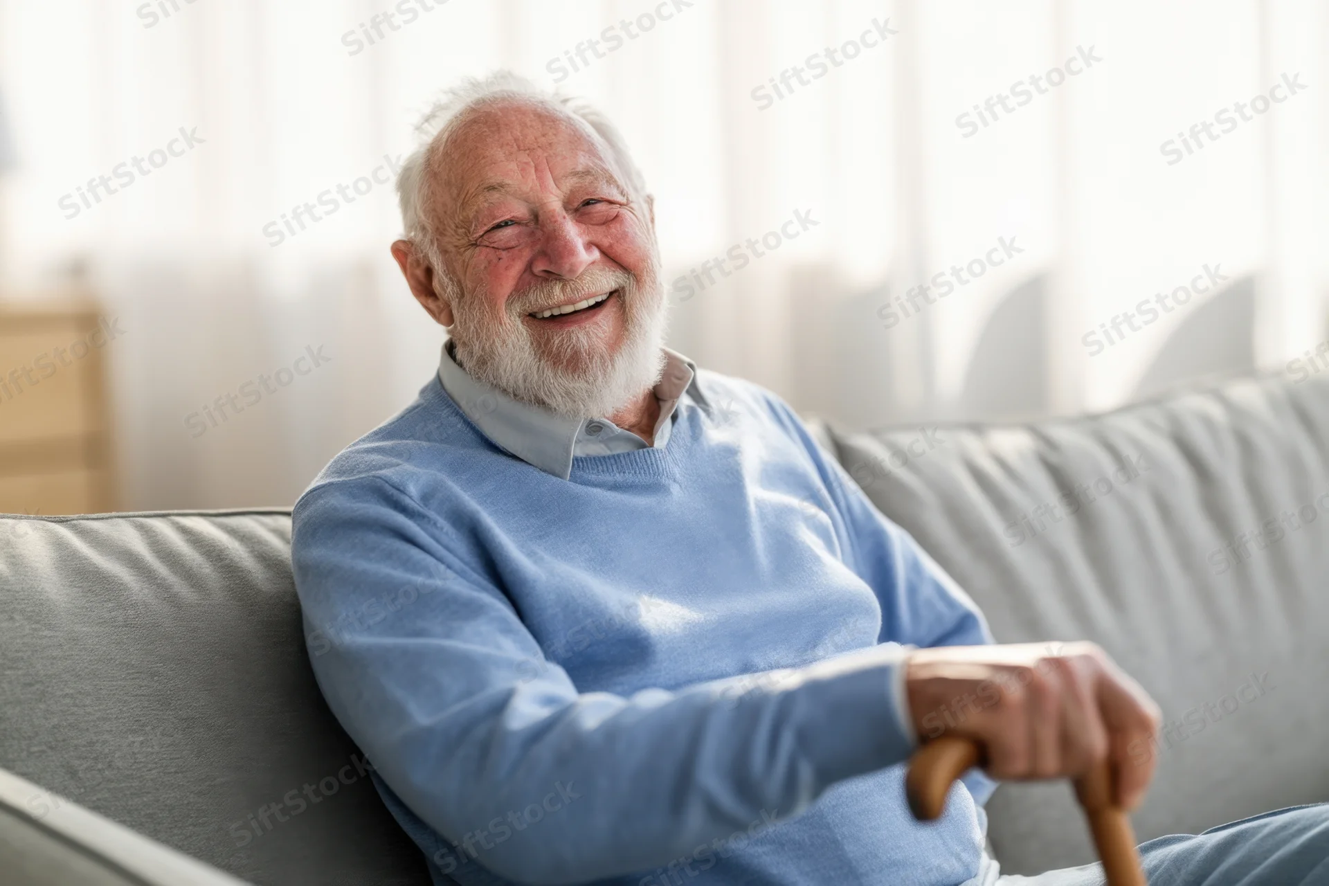 Elderly man laughing while sitting on a couch holding a wooden cane in a living room